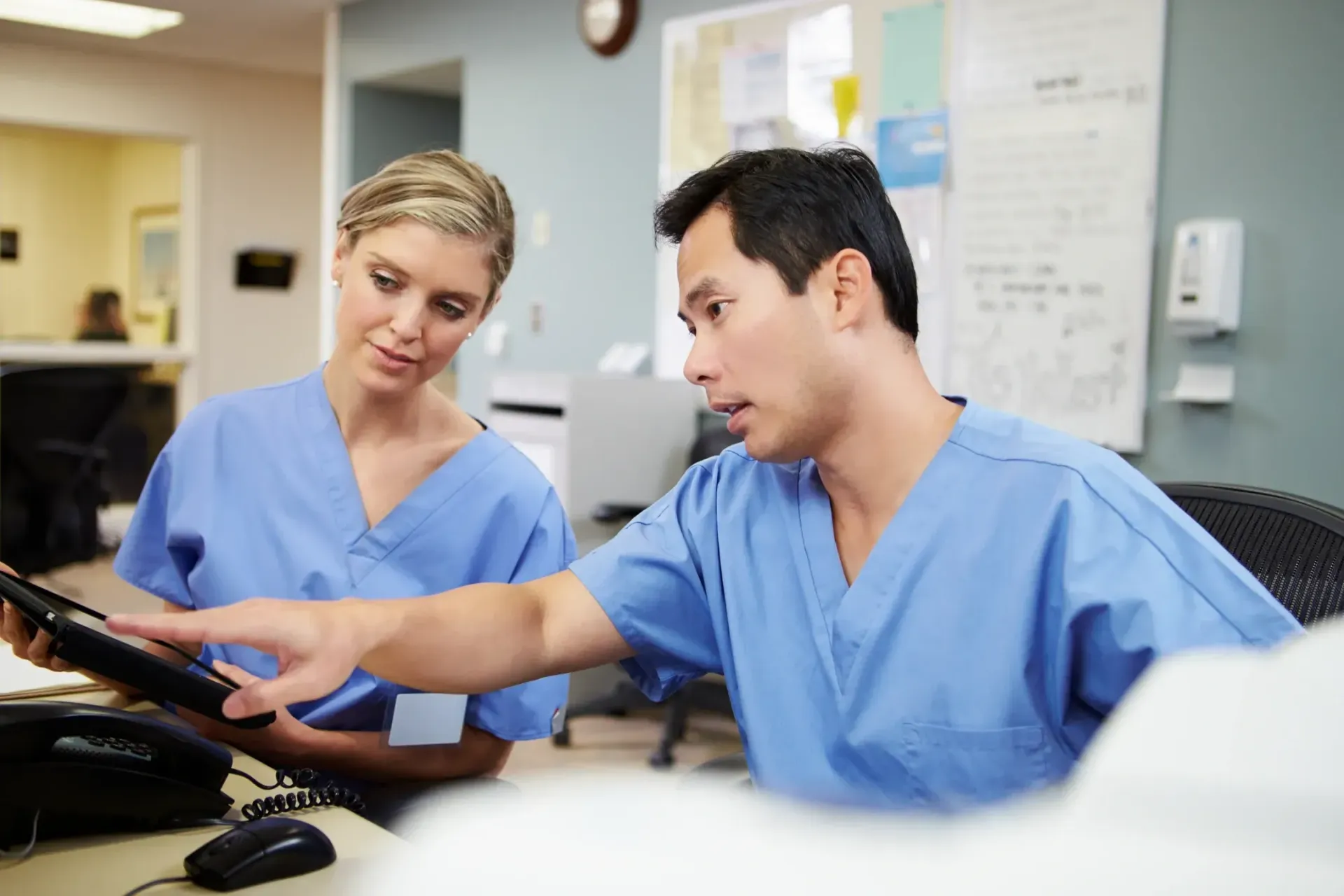 Two healthcare workers in blue scrubs reviewing a computer screen in a clinical office