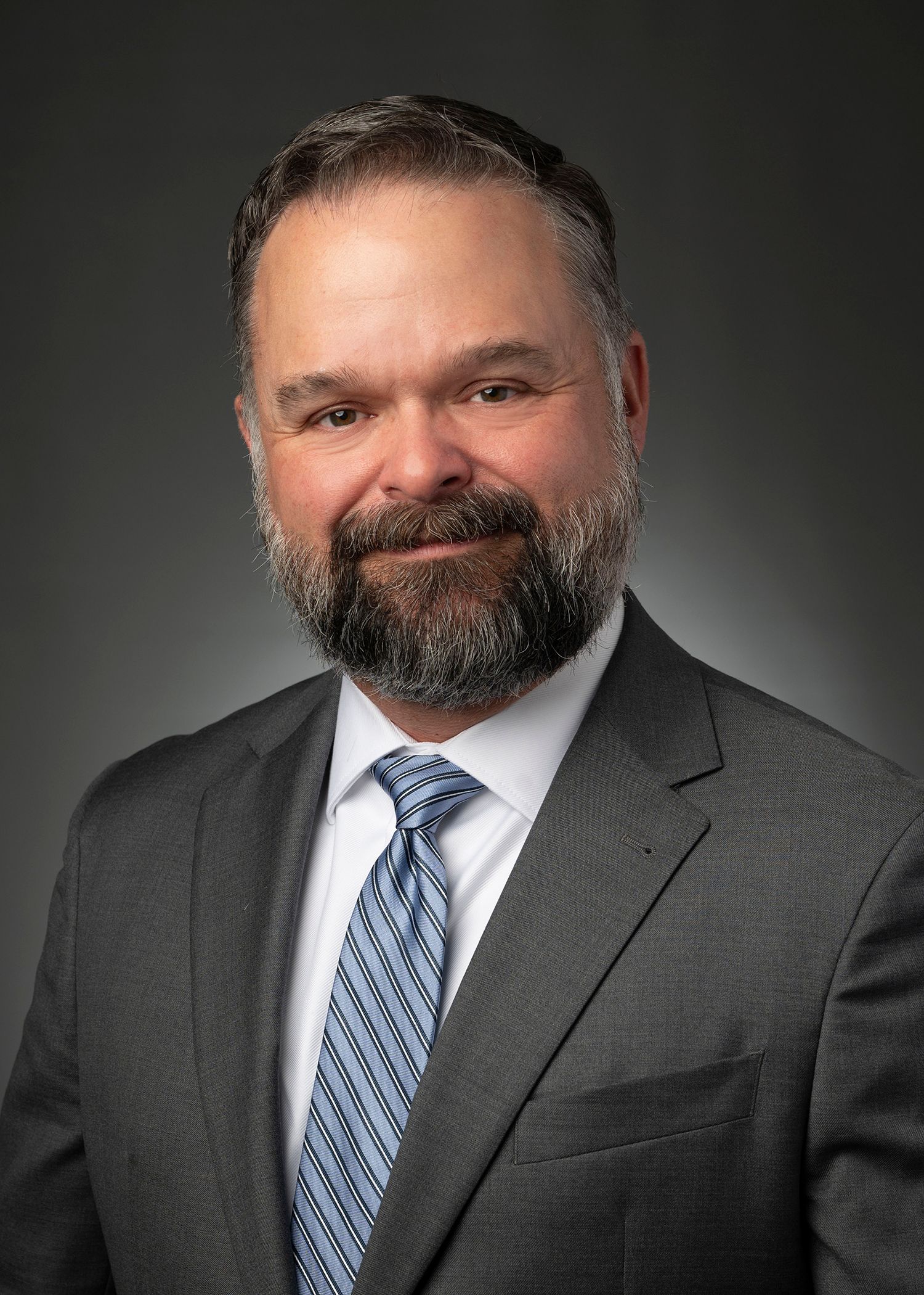 Studio portrait of a smiling man in a dark suit, white shirt, and patterned blue tie on a gray background