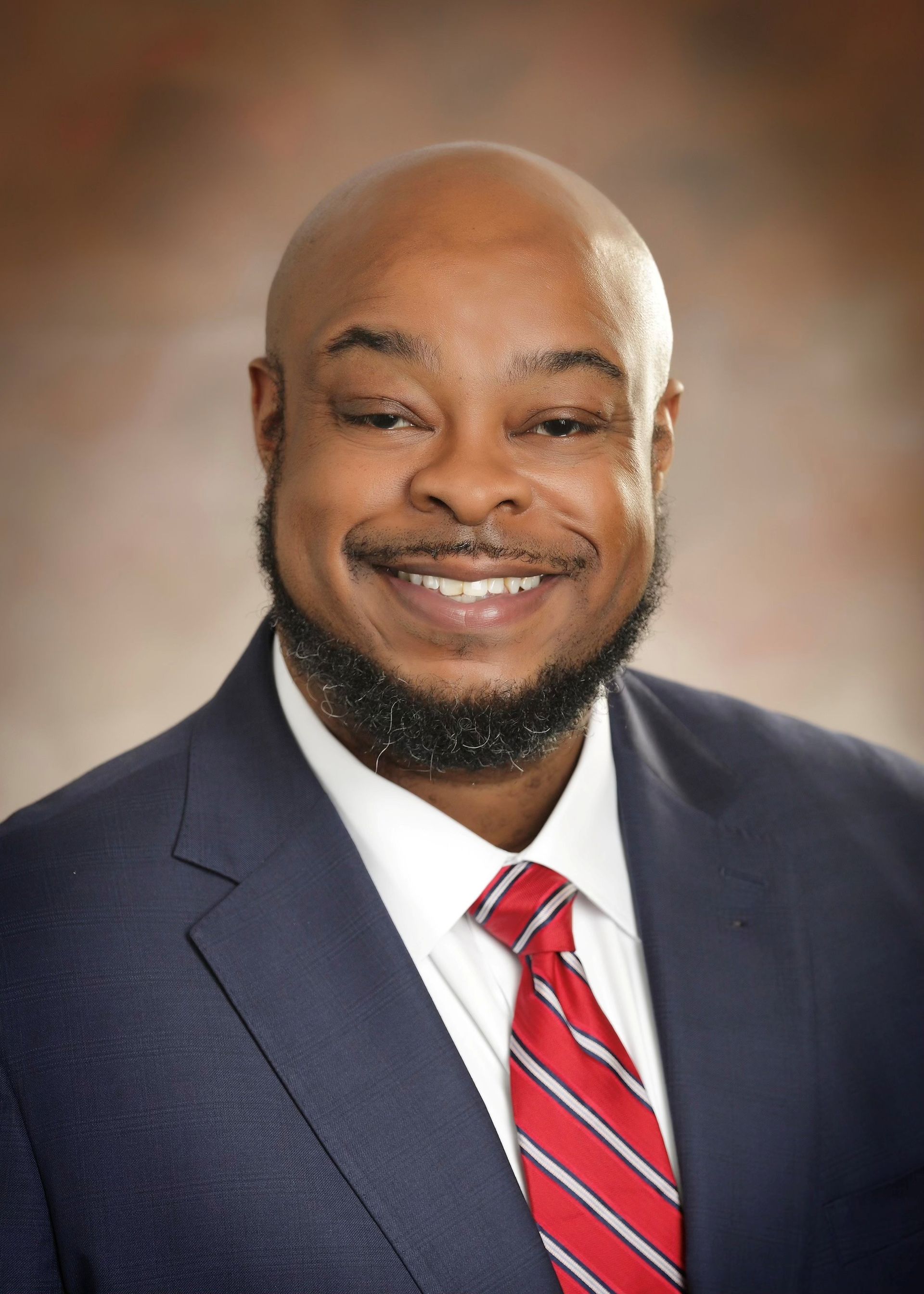 Smiling man in a navy suit and red striped tie against a soft blurred background