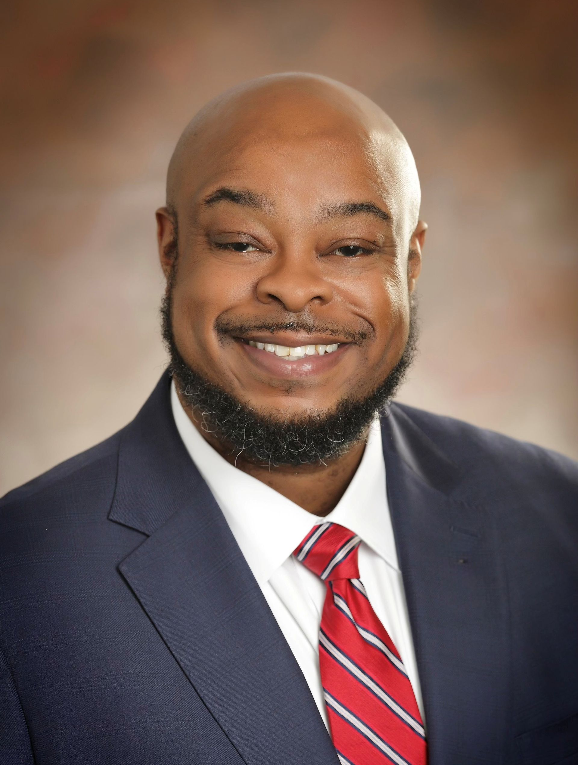 Smiling man in navy suit and red striped tie against a blurred studio background