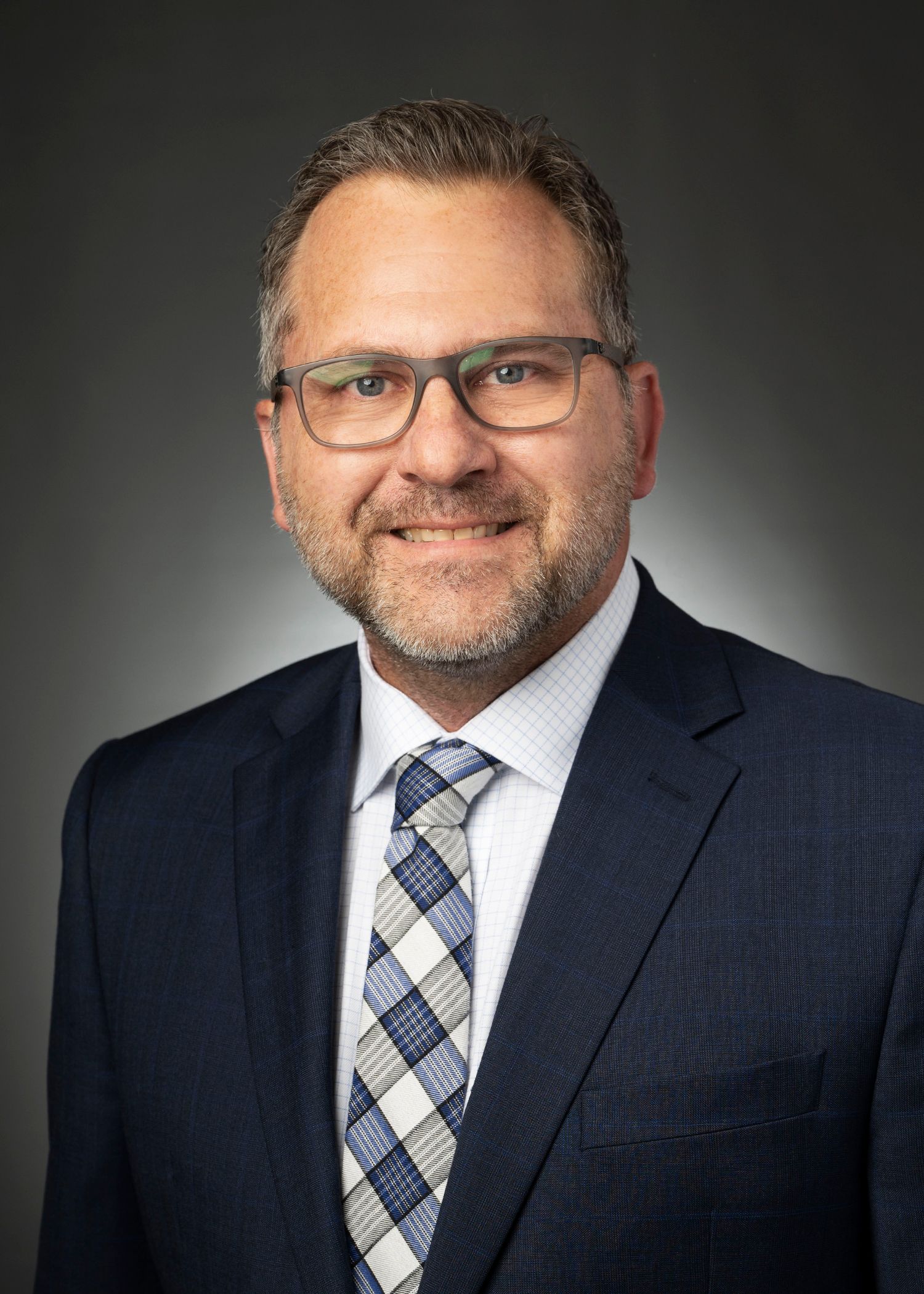 Professional headshot of a man in a navy suit and patterned tie against a dark gray background