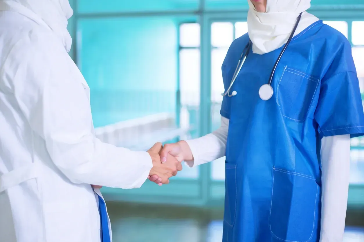 Two healthcare workers in white and blue scrubs shaking hands in a hospital hallway