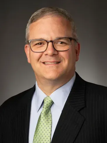 Headshot of a smiling man in glasses wearing a dark suit, light blue shirt, and green tie.