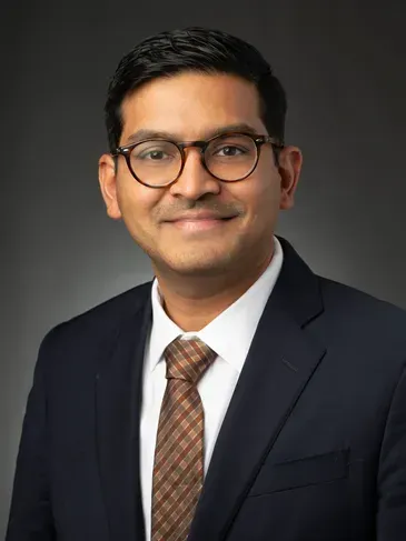 Professional headshot of a smiling person in a dark suit, white shirt, and patterned tie against a gray background