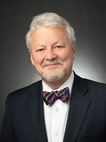 Portrait of a smiling man in a dark suit, white shirt, and colorful bow tie on a gray background