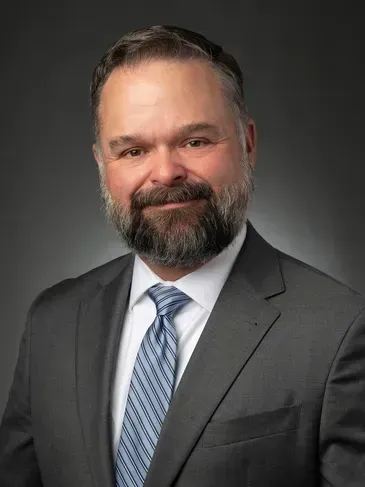 Studio headshot of a man in a gray suit and blue tie, smiling against a dark background