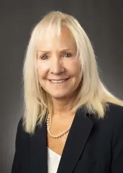 Smiling woman in a black blazer and pearl necklace against a gray studio backdrop