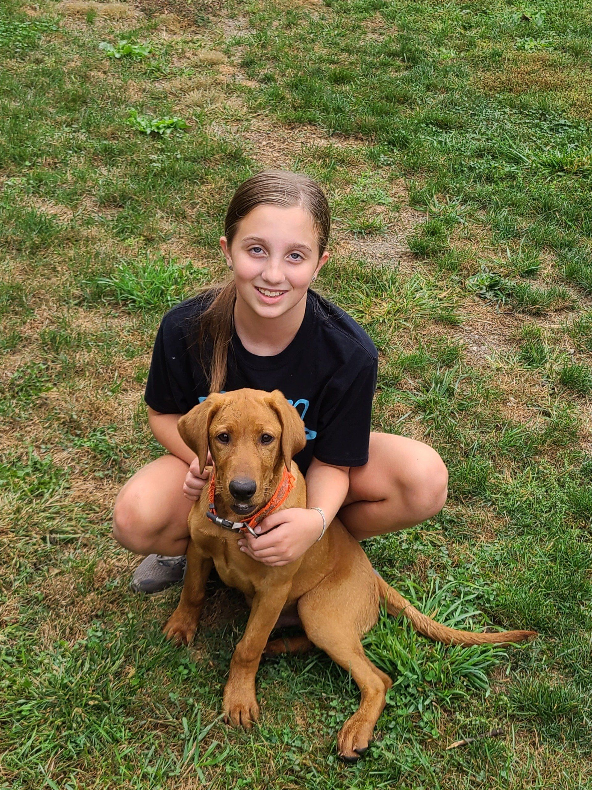 Girl crouching on grass beside a brown dog, both facing the camera.