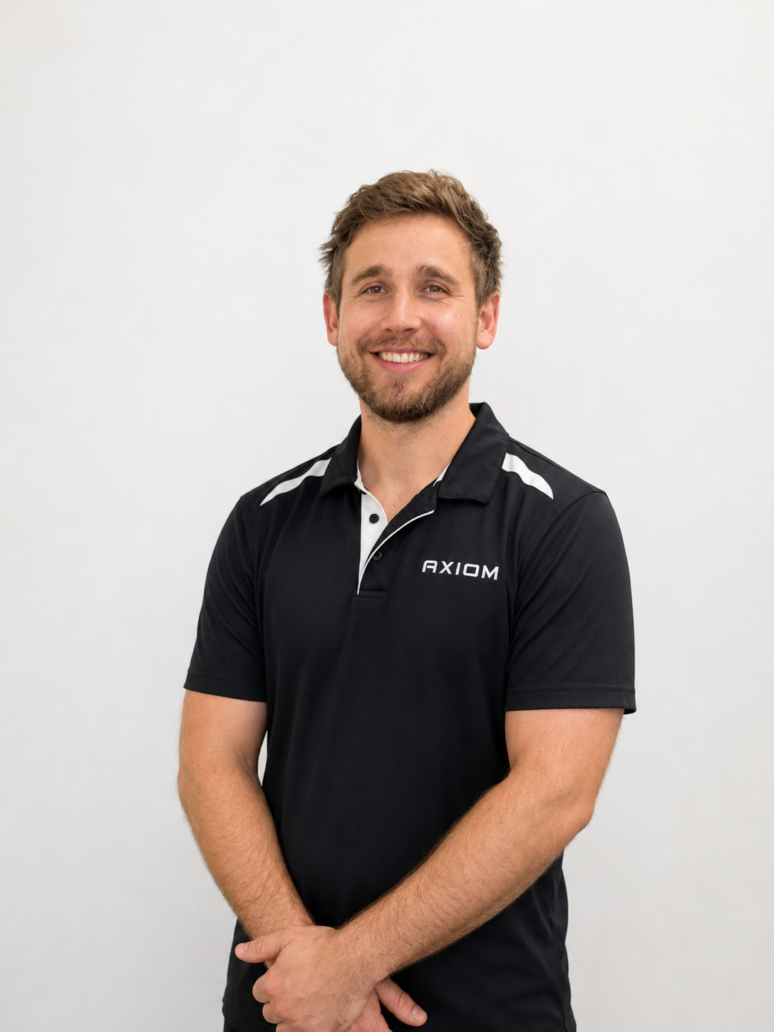 Male Remedial Massage Therapist smiles, hands crossed at waist, wearing a black and white polo shirt 