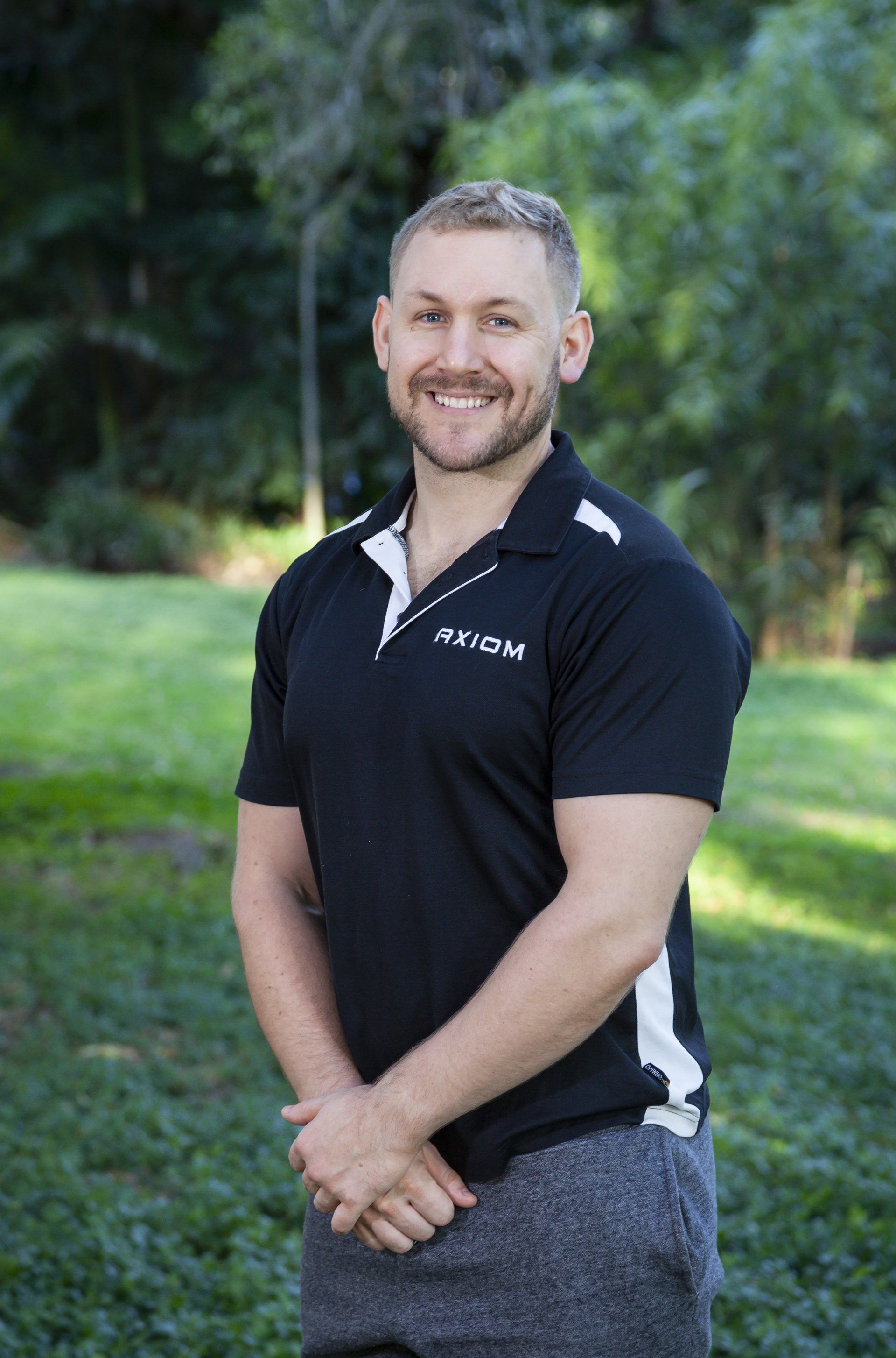 Male Remedial Massage Therapist smiling outdoors, wearing black polo shirt with white accents, arms crossed.