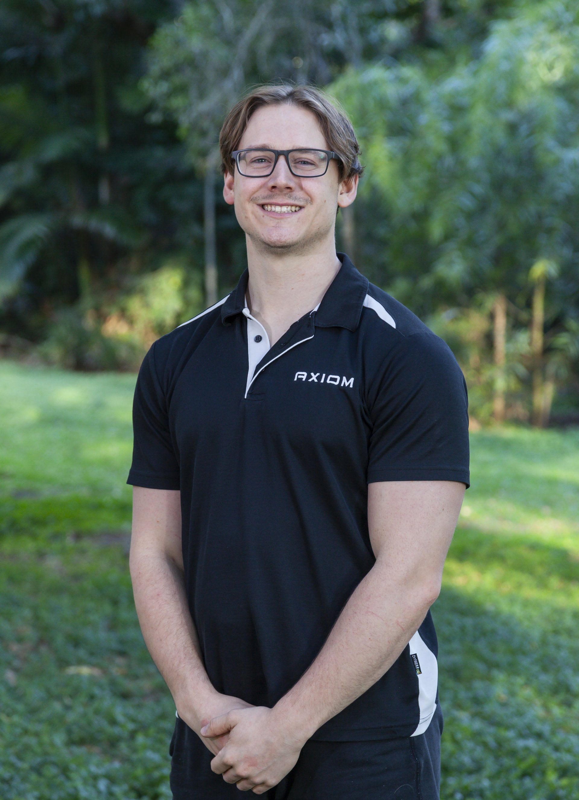 Male Remedial Massage Therapist  in glasses smiles, arms crossed, wearing a black and white polo shirt outdoors in front of greenery.
