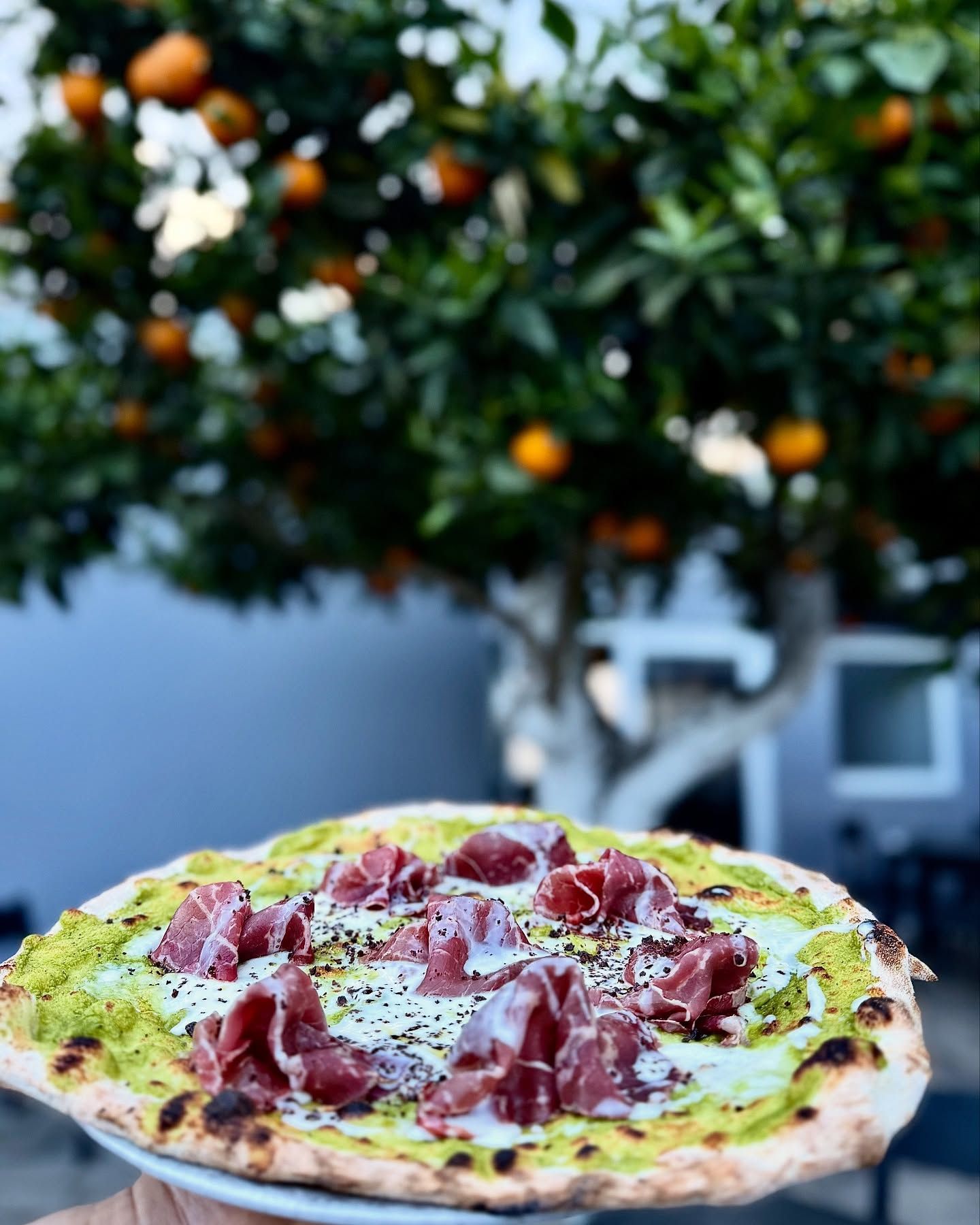 Pizza con condimento verde e prosciutto su un tavolo all'aperto sfocato, con un albero d'arancio sullo sfondo.