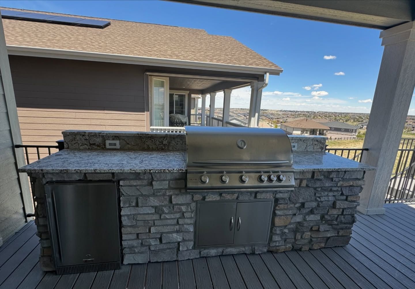 Outdoor built-in grill and countertop with stone veneer, on a deck with a house in the background.