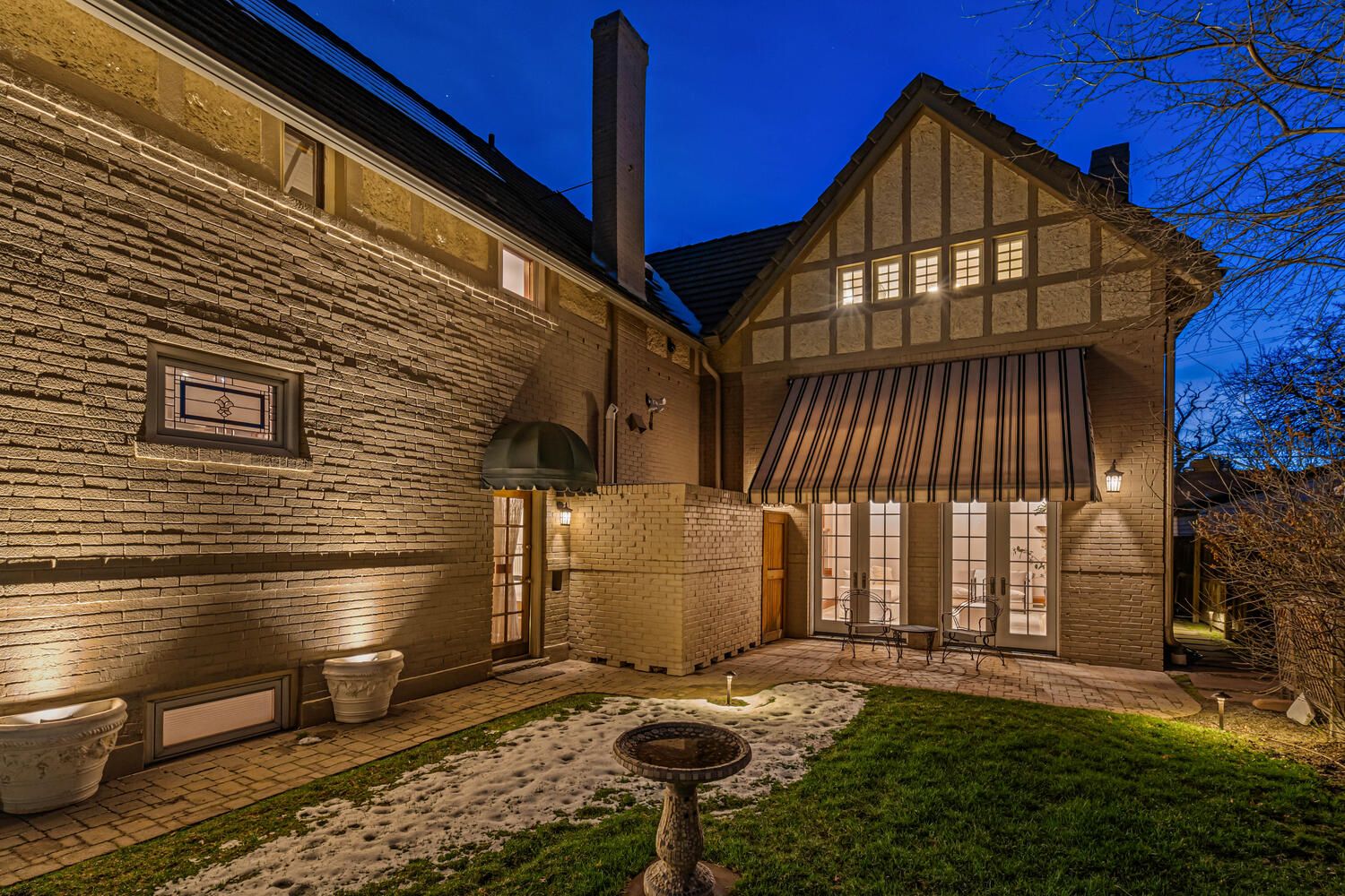 Night view of a brick house with Tudor-style gables. Lawn with snow, birdbath, and outdoor lighting.