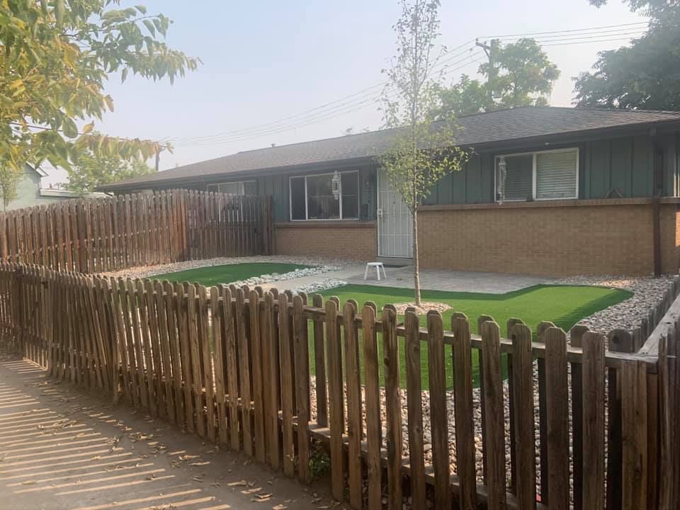 A one-story house with a wooden fence and artificial grass lawn. The house has a green and brick exterior.