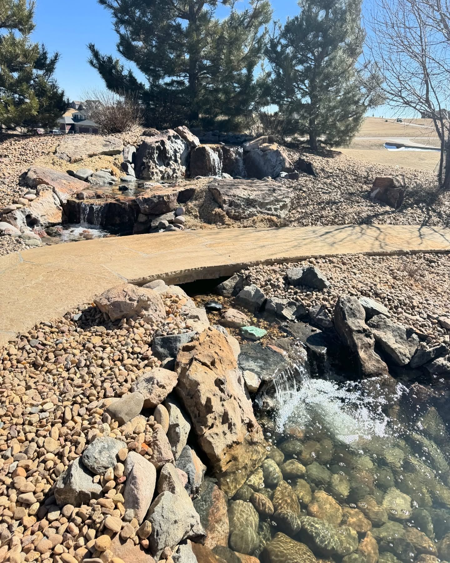 Water cascading over rocks in a landscaped setting with a walkway and trees under a clear sky.