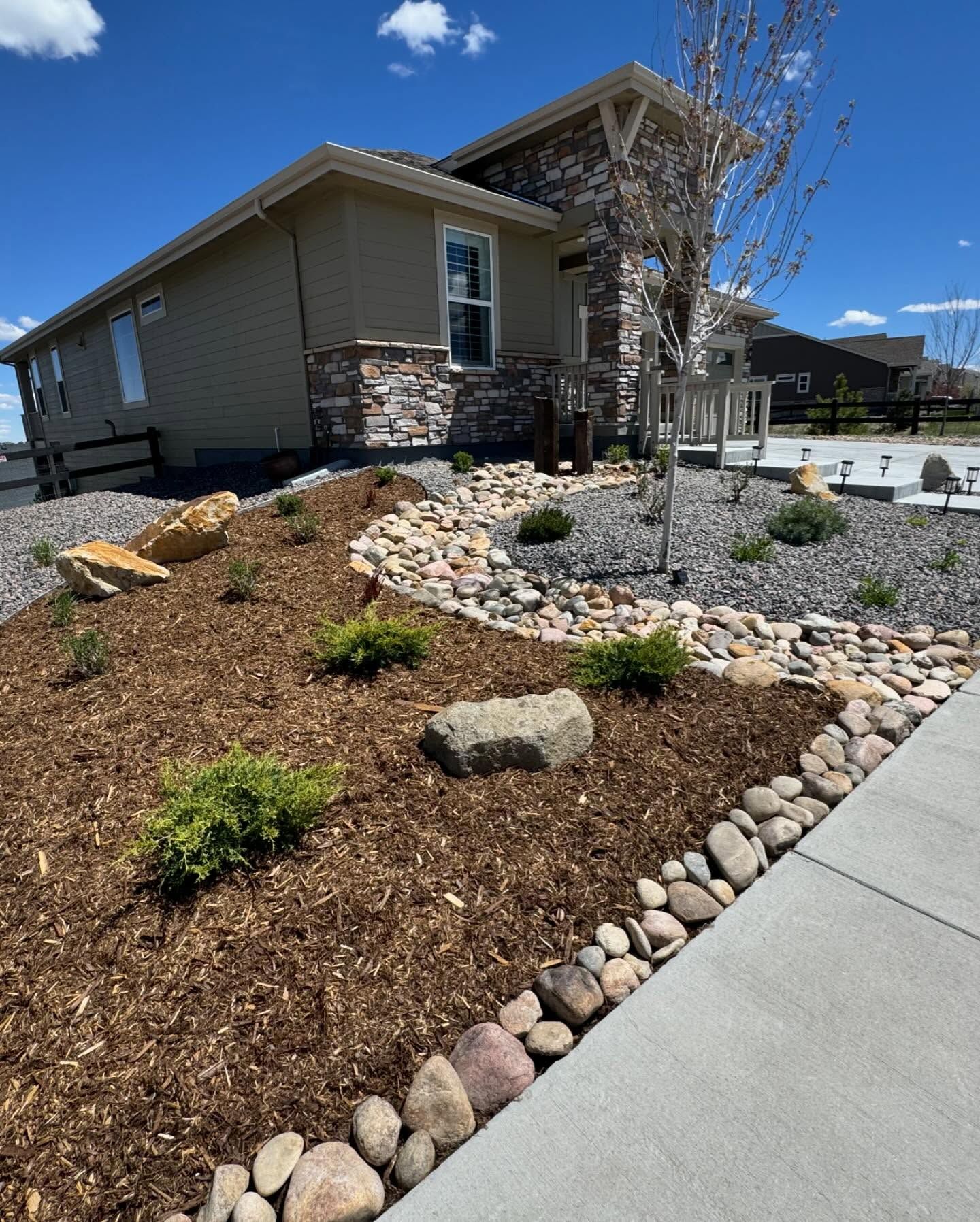 A house with landscaped front yard featuring wood chips, rocks, and small plants on a sunny day.
