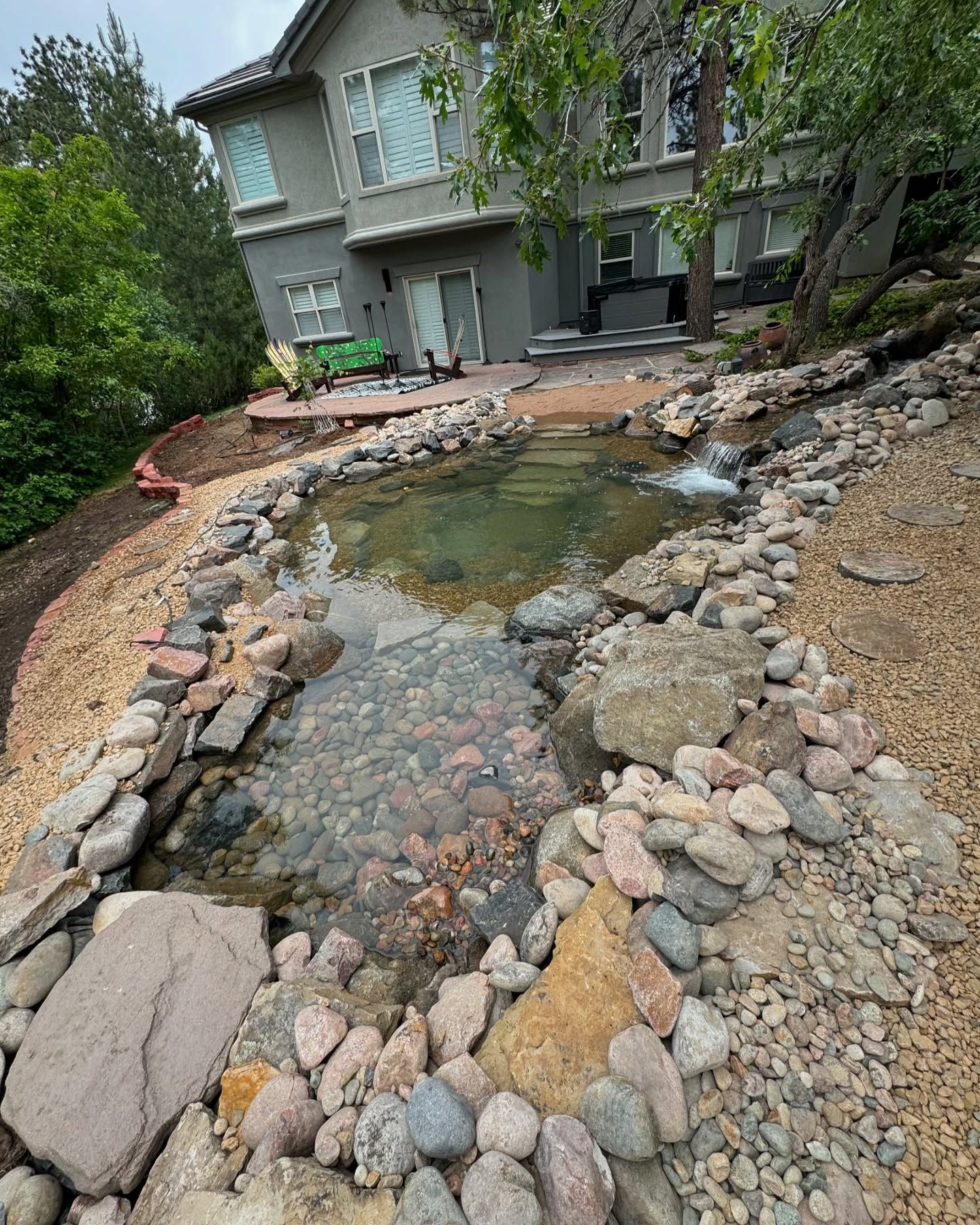 Backyard pond with waterfall, rocky edges, gravel path, and gray house in the background.