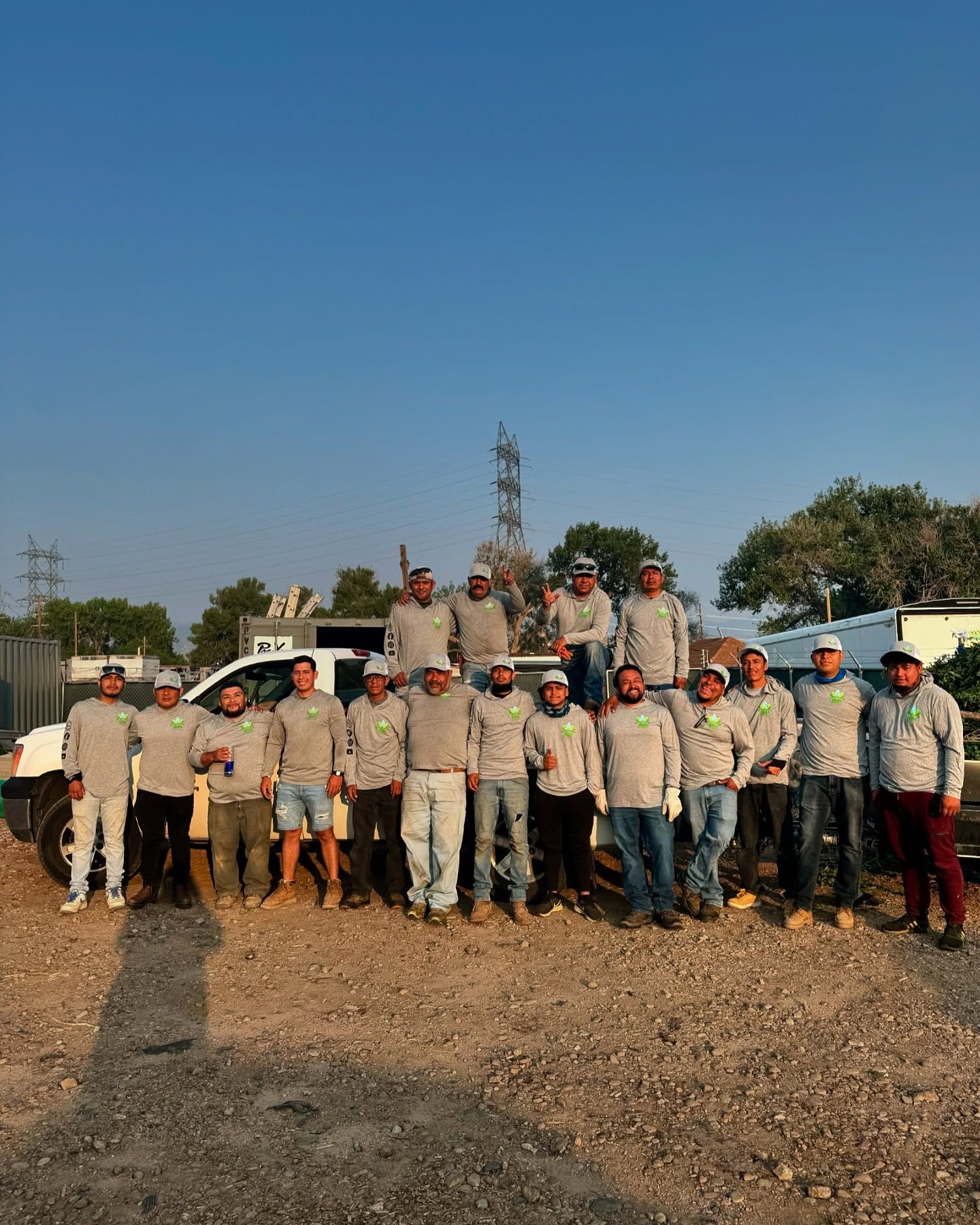 Group of workers in gray shirts and hats posing by a white truck in a dirt lot under a blue sky.