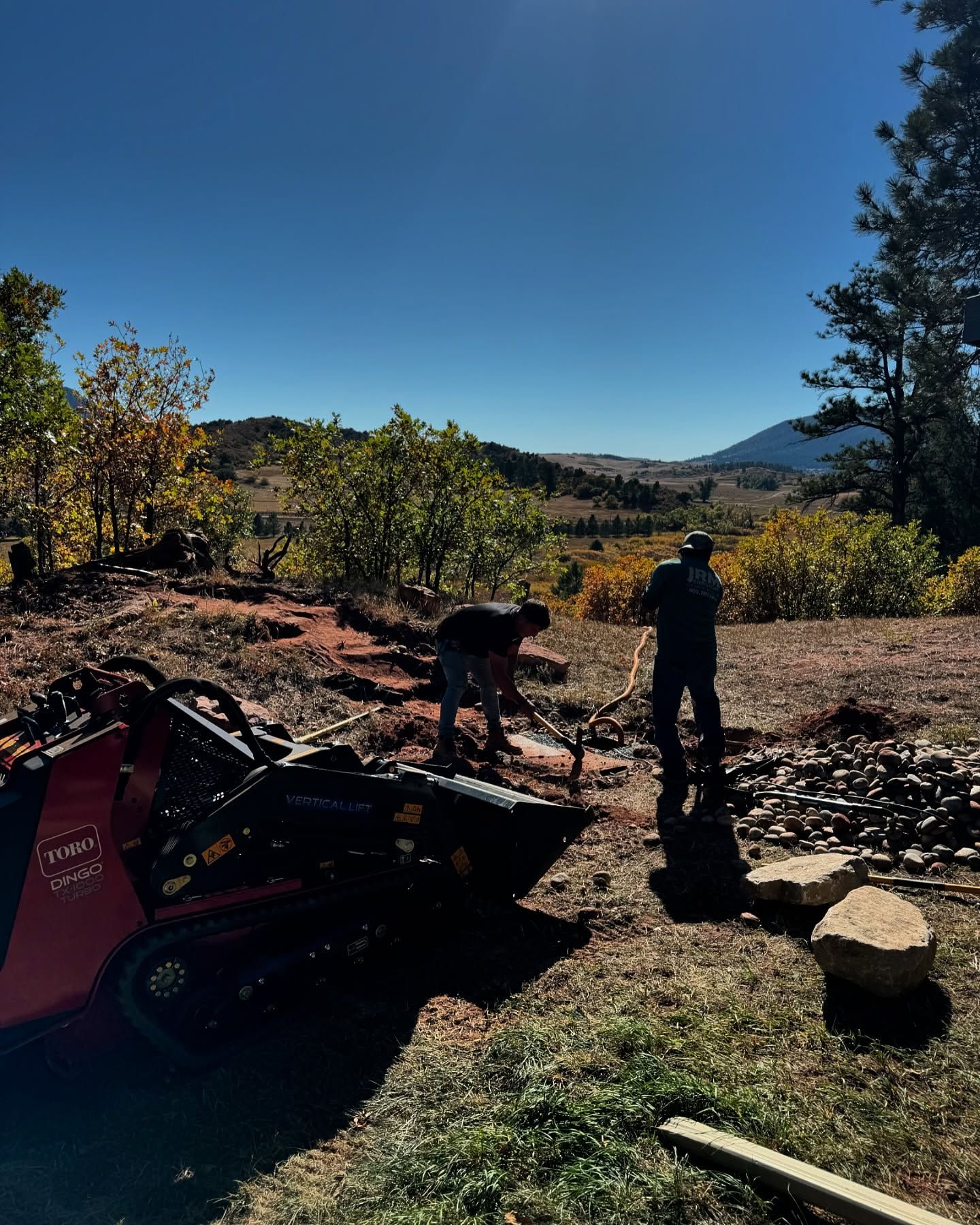 Two workers building with rocks, mini skid steer nearby, scenic view of hills and fall foliage.