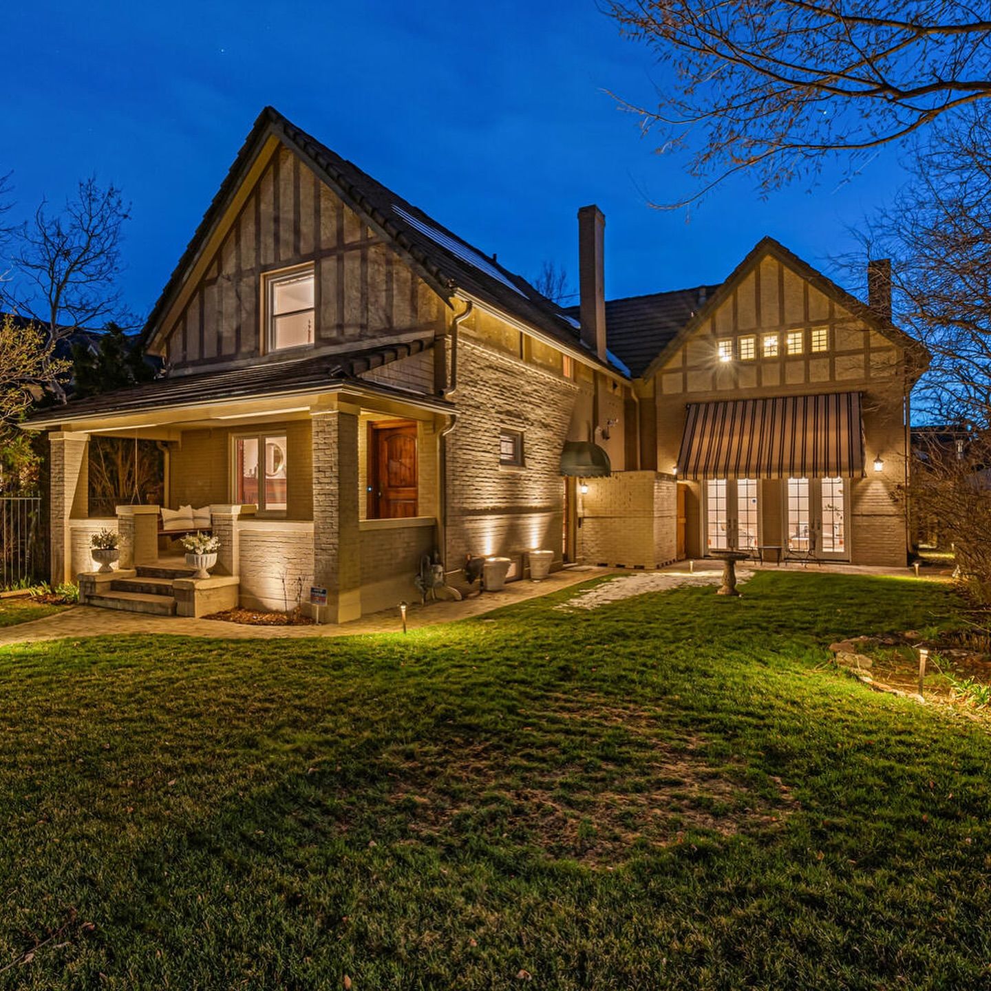 A two-story house with illuminated landscaping at dusk. Stone facade, gabled roof, and a green lawn.
