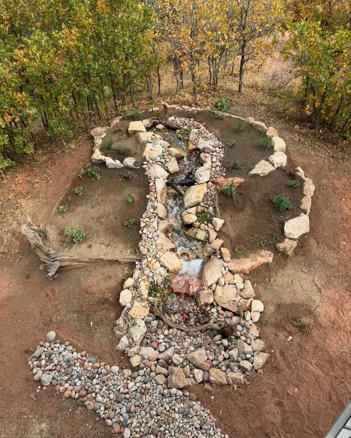 Small rock waterfall in a garden bed with surrounding trees, featuring dirt, stones, and plants.