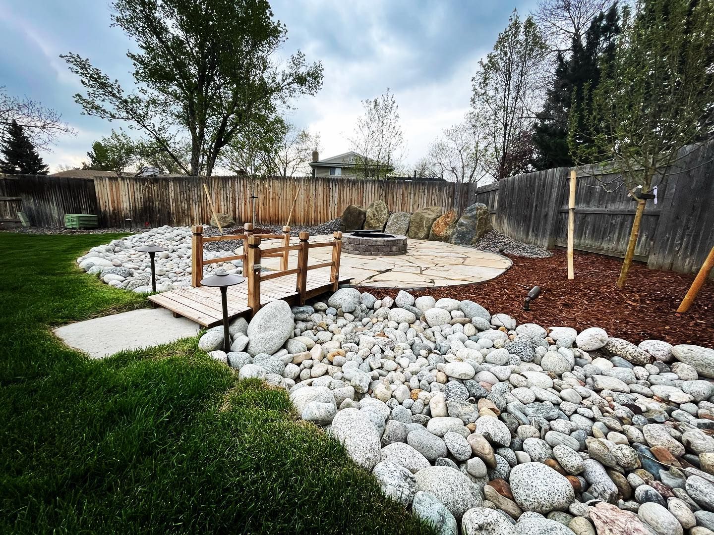 Backyard with a gravel area bordered by wood and stone, brown grass, trees, and a wooden fence.