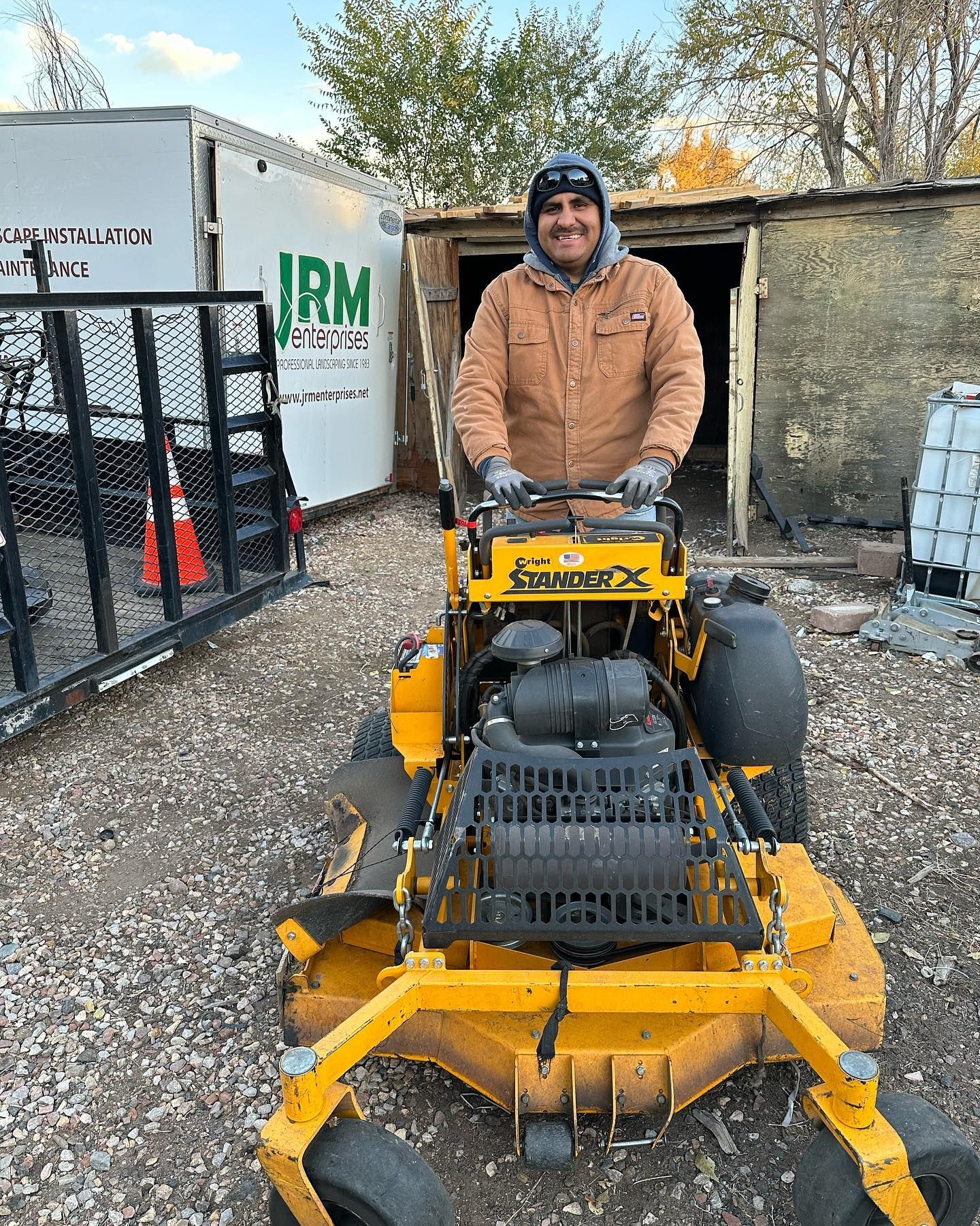 Man operating yellow lawnmower, wearing brown jacket and hat, near trailer and shed.