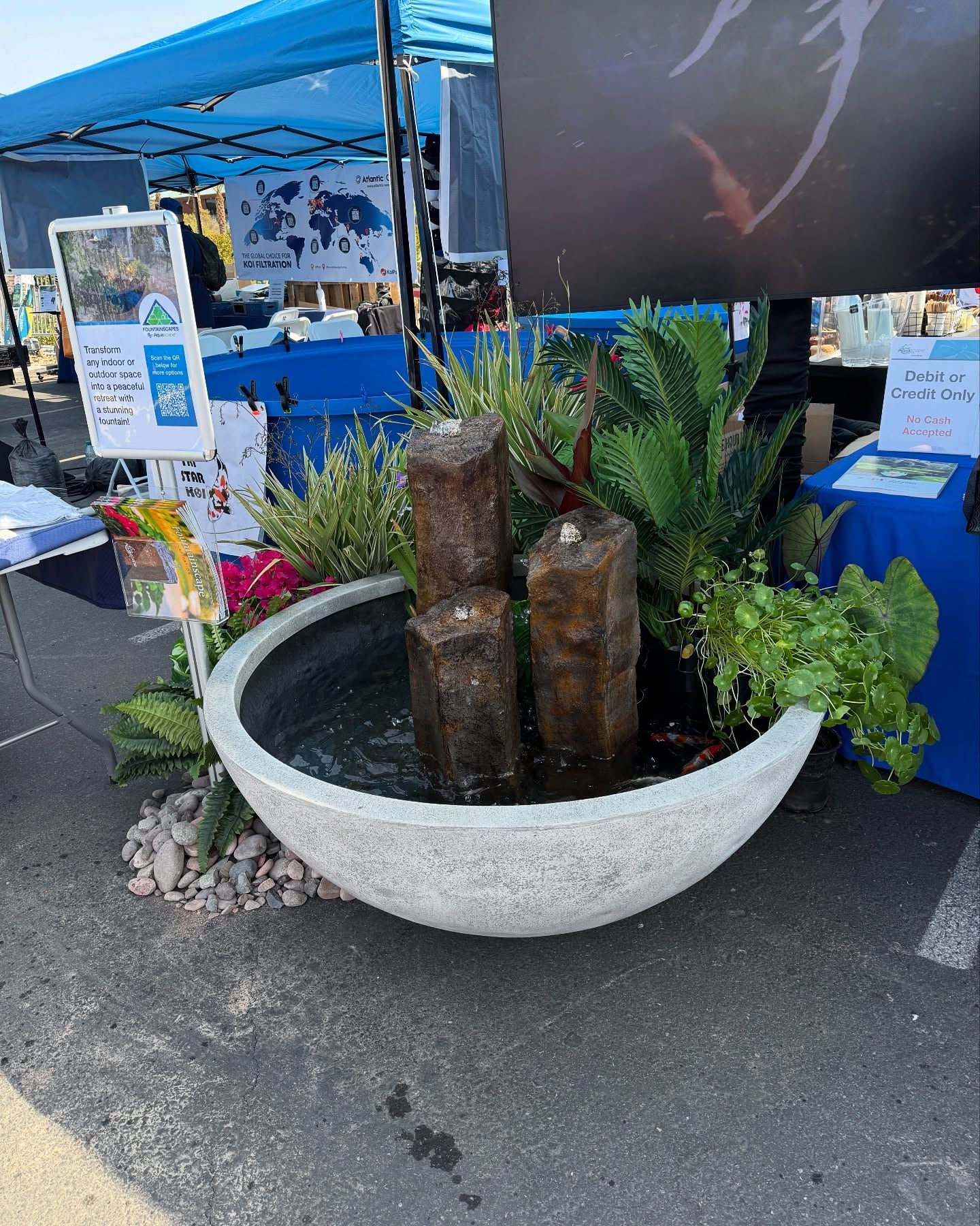 Fountain with three dark columns in a large gray bowl, surrounded by plants at a market booth.