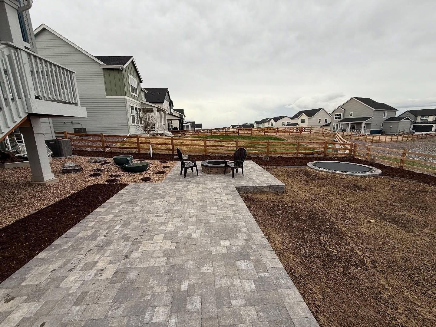 Backyard patio with paved area, fire pit, and two chairs, next to houses under an overcast sky.