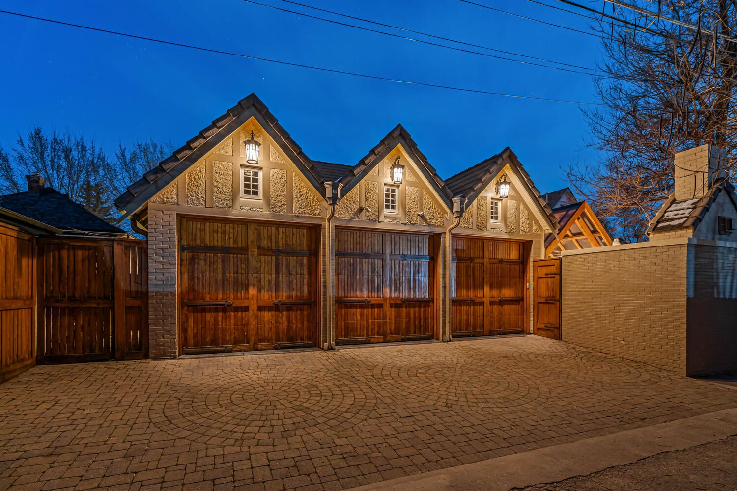 Three wooden garage doors under gabled roofs with cobblestone driveway at dusk.