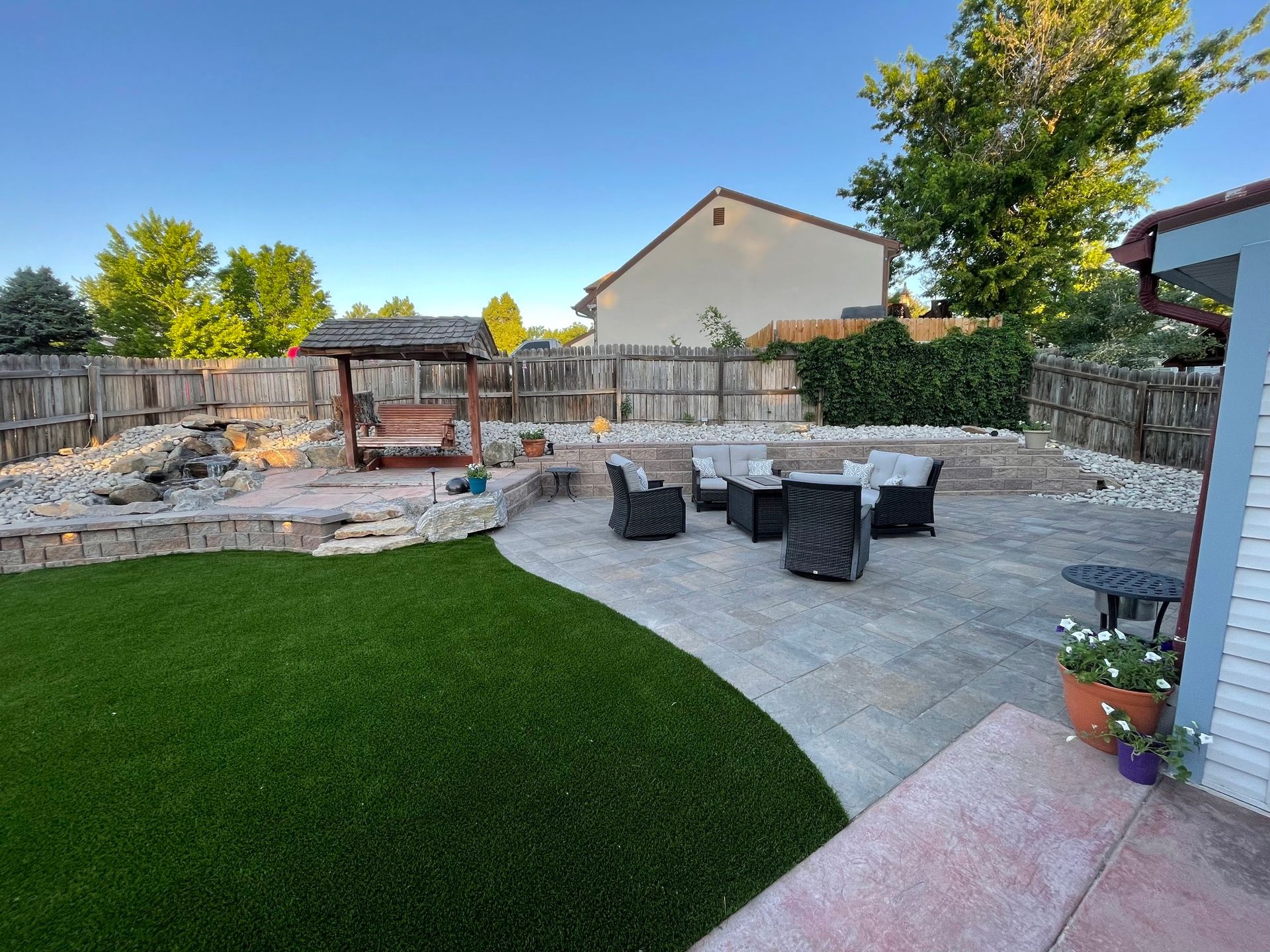 Backyard patio with stone pavers, seating, artificial grass, and a wooden swing set.
