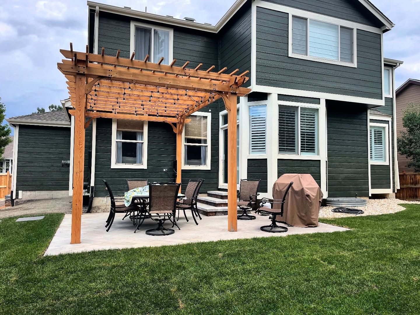 Backyard with wooden pergola over a patio, dining set, and grill; green house exterior.