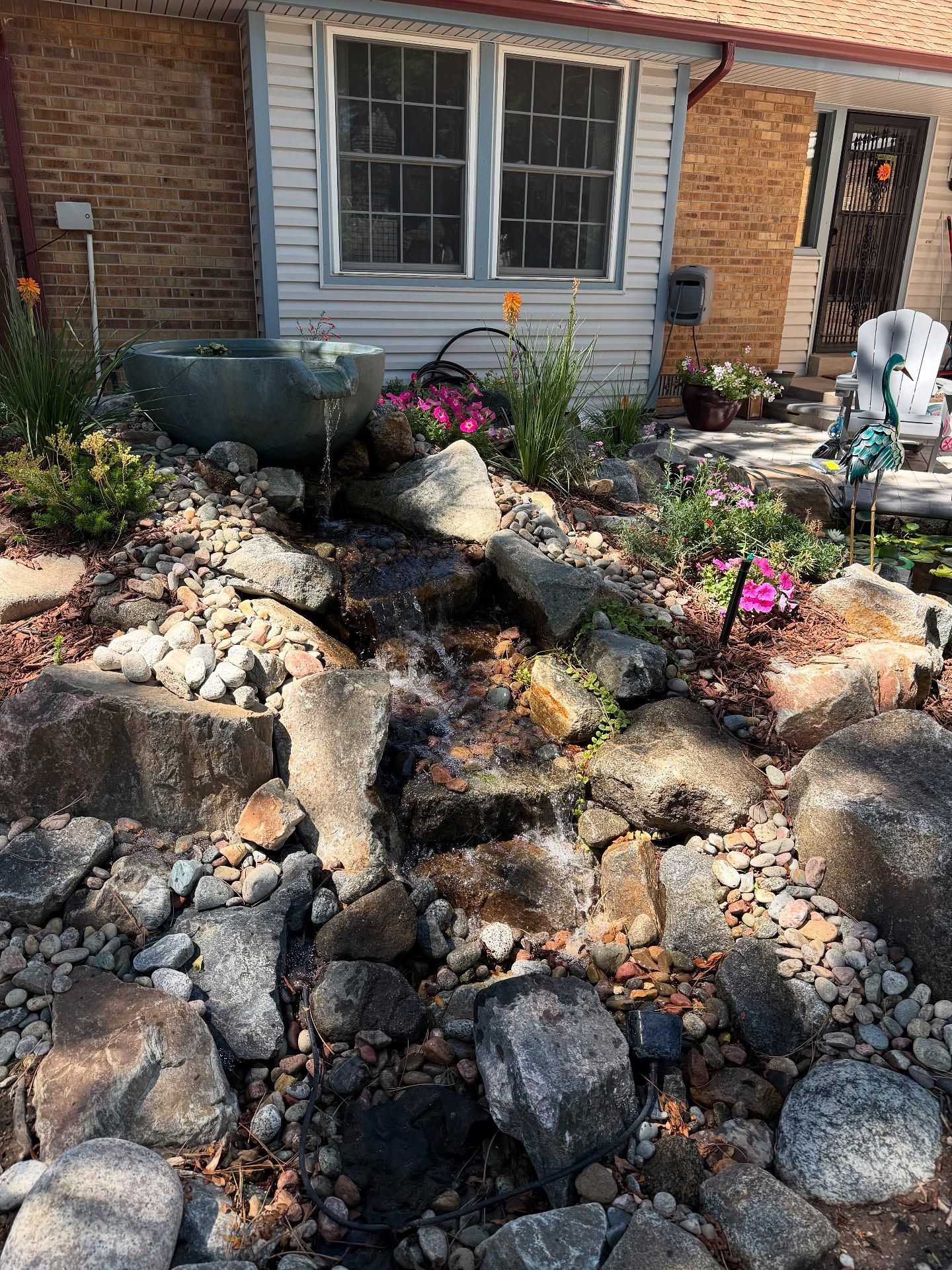 Waterfall feature cascading down rocks, leading from a basin with flowers, in a sunny garden.