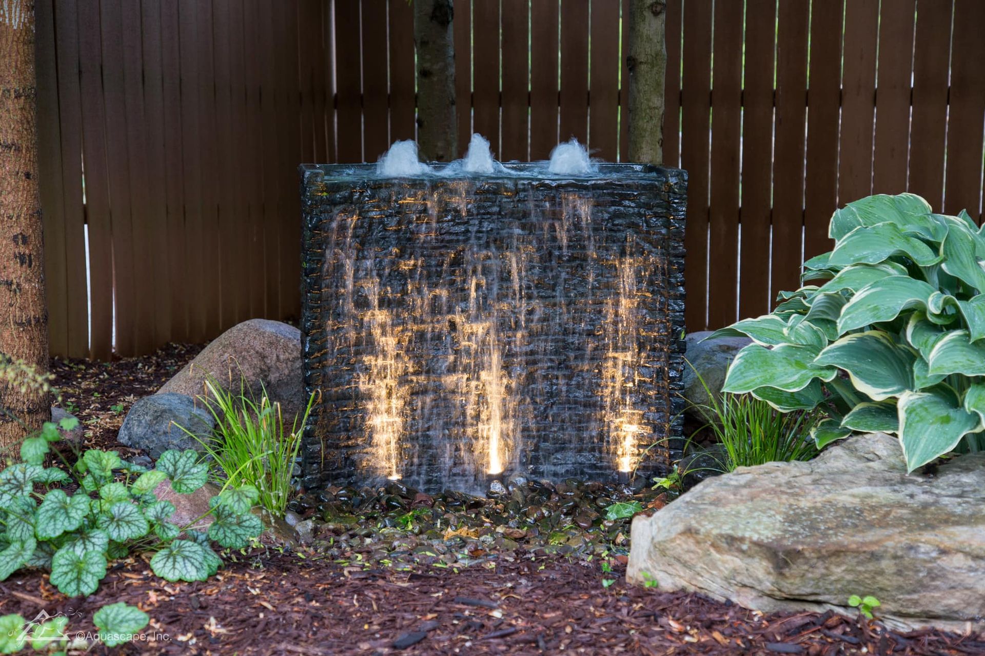 Water feature with cascading water, illuminated by three lights, in a garden setting with rocks and plants.