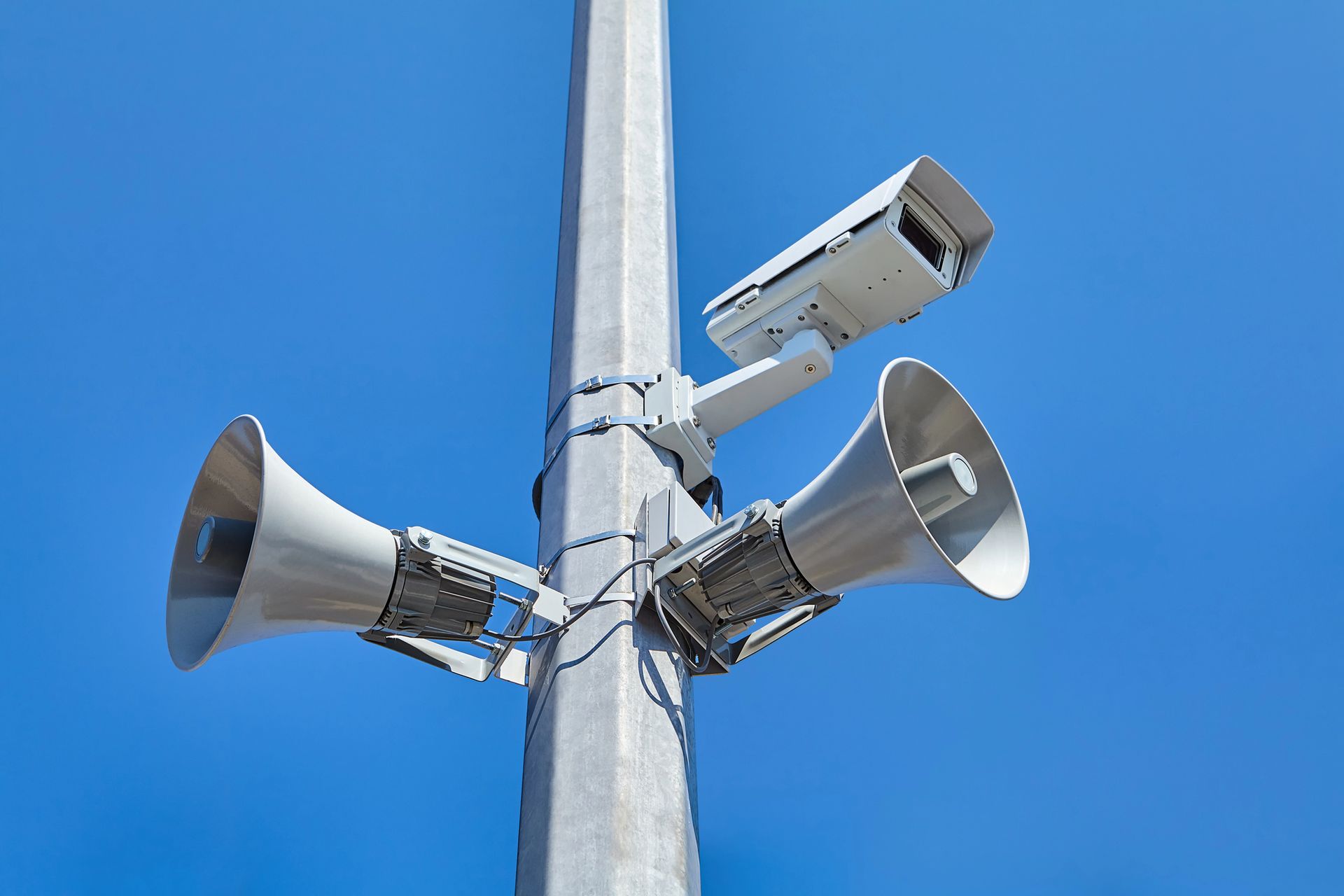Gray pole with security camera and two loudspeakers against a blue sky.