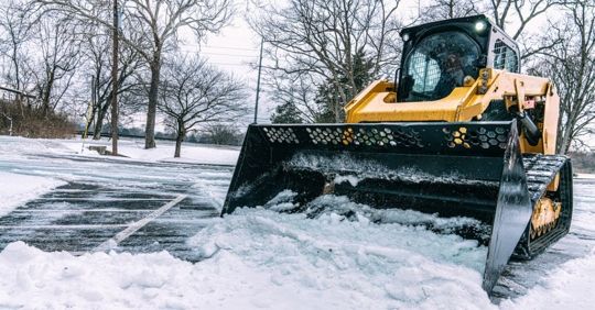 Yellow snowplow clearing snow from a parking lot on a cloudy day.