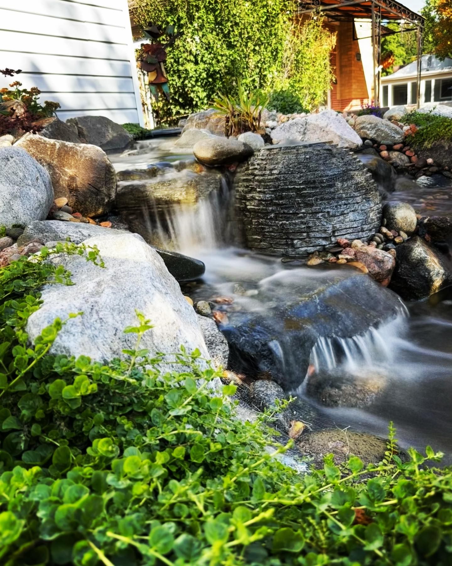 Small waterfall cascading over rocks in a garden, surrounded by lush green plants.