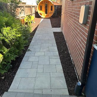 Stone pathway in backyard, leading to a wooden sauna. Brick wall and greenery on sides.