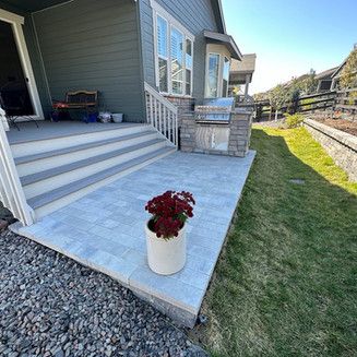 Patio with gray pavers, built-in grill, and potted flowers; green grass and house exterior in background.