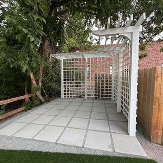 A white pergola with latticework sits on a stone patio, with a wooden fence and tree in the background.