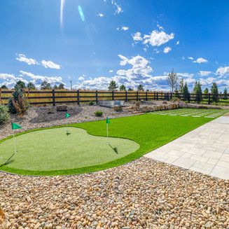 Backyard with a putting green, flagsticks, gravel, and a stone patio under a blue sky.