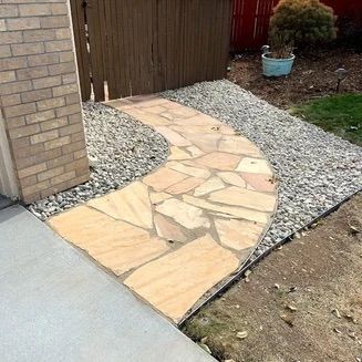 Flagstone pathway curves through gray gravel beside a brick building and wooden fence.