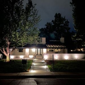 House exterior at night, illuminated by landscape lighting. White walls, dark roof, trees, and walkway.