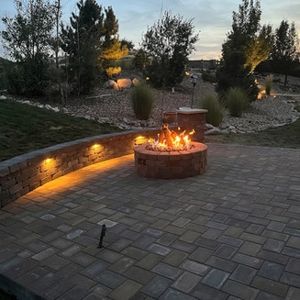 Brick patio with fire pit, glowing wall lights, and surrounding landscaping under a dusky sky.