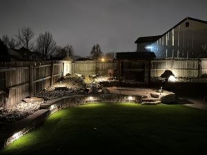 Nighttime view of a backyard with landscape lighting illuminating the lawn, stone walls, and wooden fence.