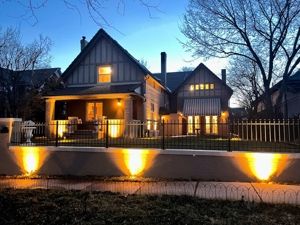 House exterior lit at night with warm lights, behind a fence.