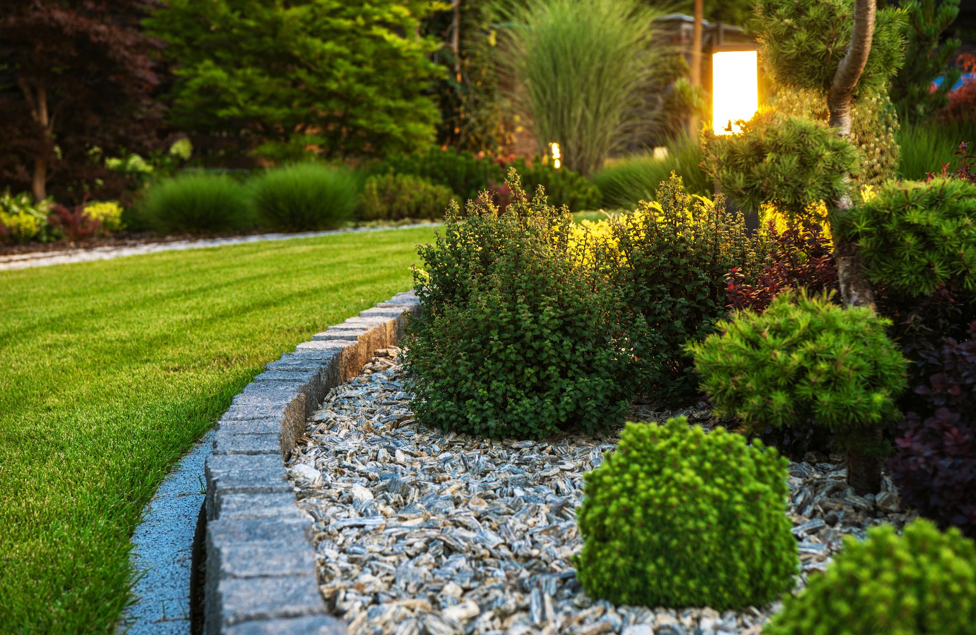 Lush garden with a stone-edged border, various green shrubs, and a grassy path leading to lit lamp.