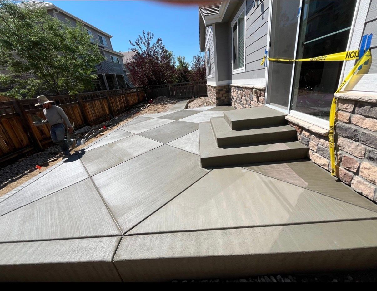 Concrete patio with steps beside a house, bordered by a wooden fence and landscaping.