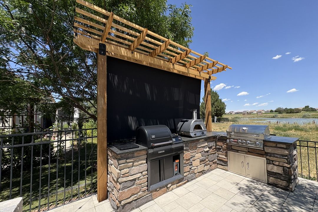 Outdoor kitchen with grill, pizza oven, and pergola, overlooking water on a sunny day.